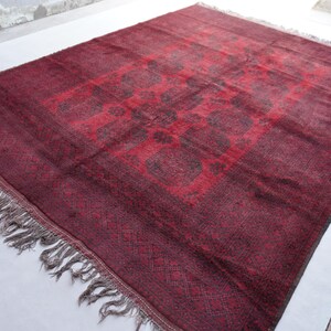 May include: A rectangular, burgundy-colored rug with a central geometric design in a darker shade of red. The rug has a fringe along the edges and a patterned border. The rug is likely made of wool or a similar material.