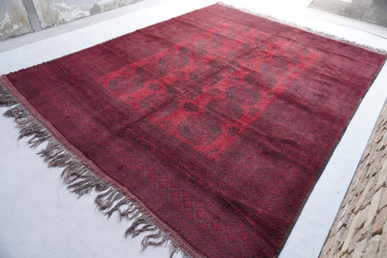 May include: A rectangular, burgundy-colored rug with a central geometric design in a darker shade of red. The rug has a fringe along the edges and a patterned border. The rug is likely made of wool or a similar material.