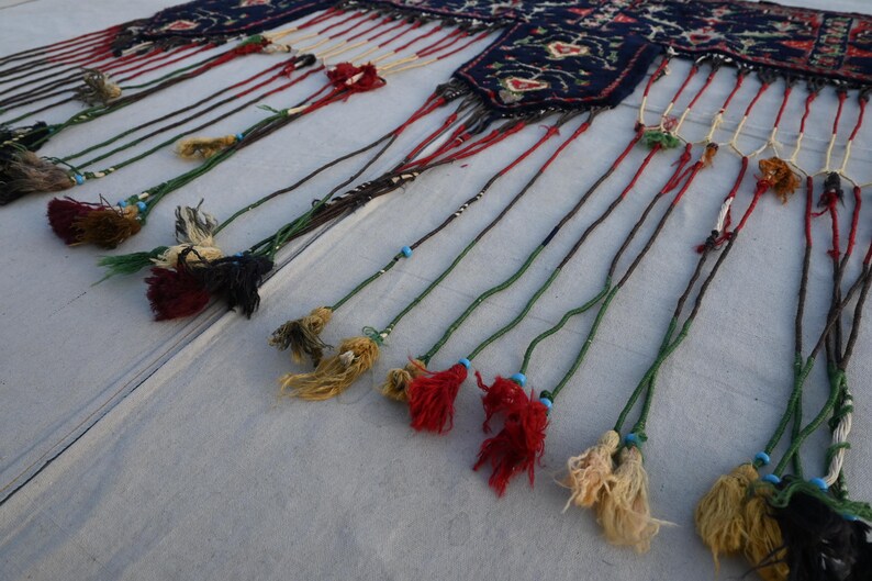 Puede incluir: Primer plano de un textil decorativo con una parte superior estampada en azul oscuro y largas y coloridas borlas. Las borlas son de varios colores, incluyendo rojo, verde y amarillo, con peque&ntilde;as cuentas. El textil se utiliza probablemente para la decoraci&oacute;n del hogar.