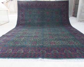May include: A large, rectangular rug with a detailed geometric pattern in shades of green, red, and blue. The rug has a dark blue border and a fringe along one edge. The design features repeating floral and geometric motifs.