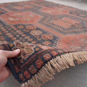 Pu&ograve; includere: Una mano solleva un angolo di un tappeto vintage, rivelando il suo intricato motivo geometrico. Il tappeto presenta una miscela di tonalit&agrave; rosso ruggine e blu navy, con un bordo sfrangiato. Il design include motivi a diamante e floreali, creando un'estetica classica e invecchiata.