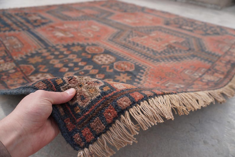 Pu&ograve; includere: Una mano solleva un angolo di un tappeto vintage, rivelando il suo intricato motivo geometrico. Il tappeto presenta una miscela di tonalit&agrave; rosso ruggine e blu navy, con un bordo sfrangiato. Il design include motivi a diamante e floreali, creando un'estetica classica e invecchiata.