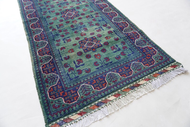 May include: A rectangular green rug with a detailed pattern. The rug features a central field of green with floral and geometric designs in blue and red. The border is a darker blue with repeating patterns and white fringe.