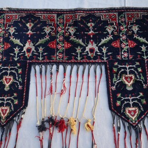 K&ouml;nnte beinhalten: Ein dunkelblaues Textil mit aufwendigen floralen und geometrischen Mustern in Rot, Wei&szlig; und Gr&uuml;n. Das Textil hat einen Fransenrand mit Quasten in verschiedenen Farben. Das Design ist symmetrisch und detailliert.