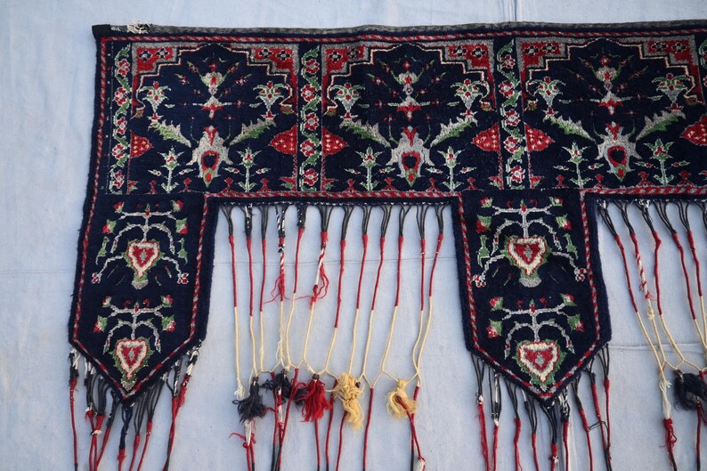K&ouml;nnte beinhalten: Ein dunkelblaues Textil mit aufwendigen floralen und geometrischen Mustern in Rot, Wei&szlig; und Gr&uuml;n. Das Textil hat einen Fransenrand mit Quasten in verschiedenen Farben. Das Design ist symmetrisch und detailliert.
