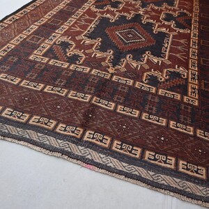 May include: A rectangular, hand-woven rug with a geometric pattern. The rug features a central diamond design in dark brown and black, surrounded by repeating patterns in beige and brown. The border is a darker shade of brown with geometric shapes.