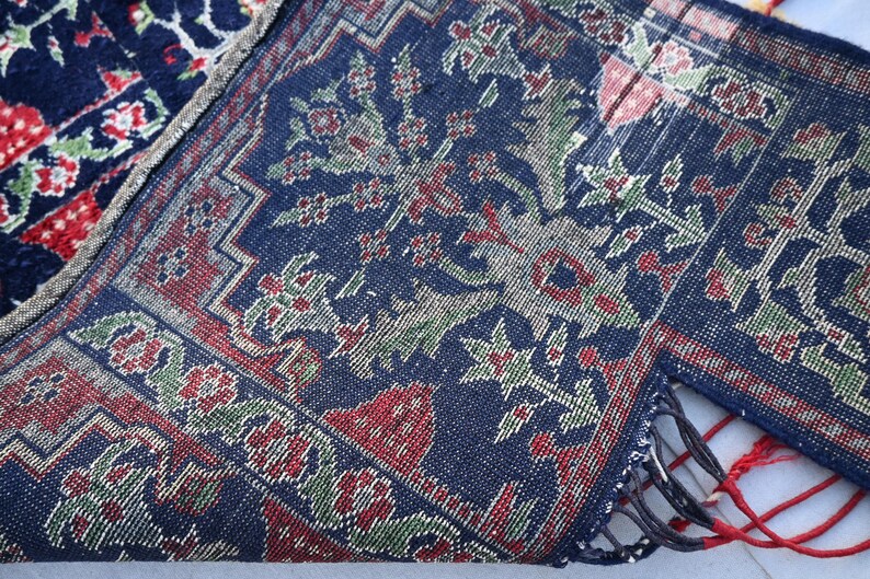 K&ouml;nnte beinhalten: Nahaufnahme eines Teils eines dunkelblauen Teppichs mit einem Blumenmuster in Rot, Gr&uuml;n und Wei&szlig;. Der Teppich hat einen detaillierten Rand und Fransen. Der Teppich ist gefaltet und zeigt die Unterseite.