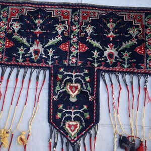 K&ouml;nnte beinhalten: Nahaufnahme eines dunkelblauen Textils mit aufwendigen Mustern in Rot, Wei&szlig; und Gr&uuml;n. Das Textil hat ein zentrales Feld mit herzf&ouml;rmigem Design und lange, dekorative Fransen in Rot, Schwarz und Gelb.