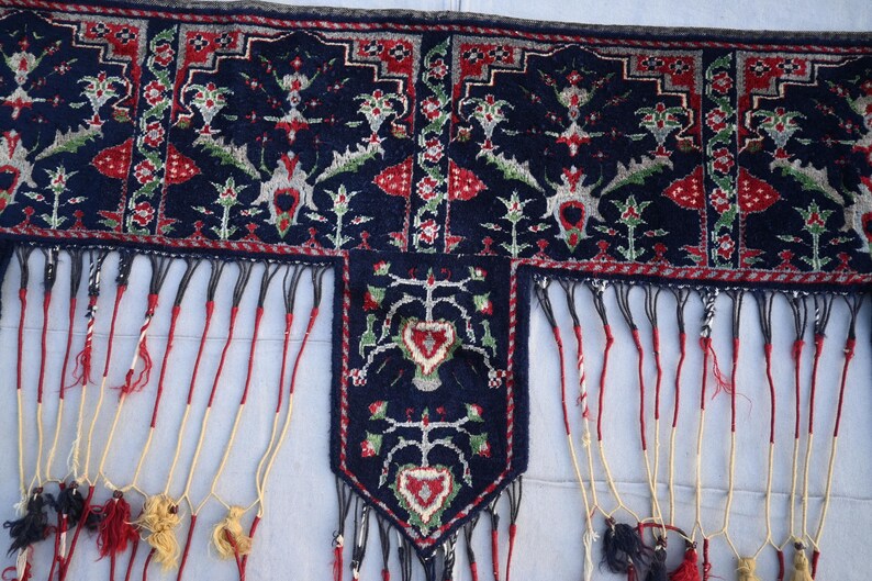 K&ouml;nnte beinhalten: Nahaufnahme eines dunkelblauen Textils mit aufwendigen Mustern in Rot, Wei&szlig; und Gr&uuml;n. Das Textil hat ein zentrales Feld mit herzf&ouml;rmigem Design und lange, dekorative Fransen in Rot, Schwarz und Gelb.