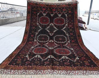 May include: A large, rectangular rug with a central pattern of red and black geometric shapes. The rug has a dark brown border and a cream-coloured fringe. The design includes diamond and octagon shapes.