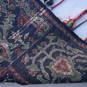Puede incluir: Primer plano de una secci&oacute;n de una alfombra azul oscuro con un dise&ntilde;o floral en rojo, verde y beige. El borde de la alfombra es visible, con borlas rojas, verdes y marrones. La alfombra tiene una textura tejida detallada.