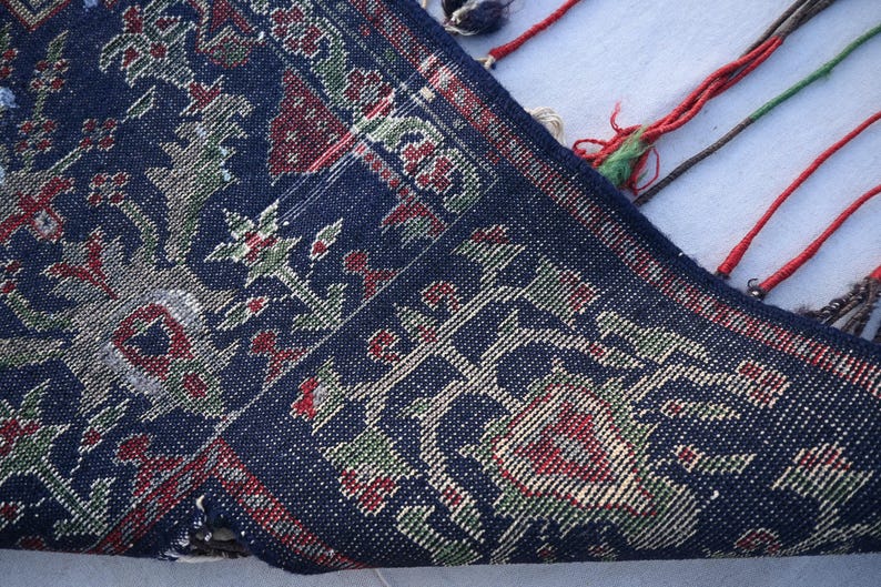 Puede incluir: Primer plano de una secci&oacute;n de una alfombra azul oscuro con un dise&ntilde;o floral en rojo, verde y beige. El borde de la alfombra es visible, con borlas rojas, verdes y marrones. La alfombra tiene una textura tejida detallada.
