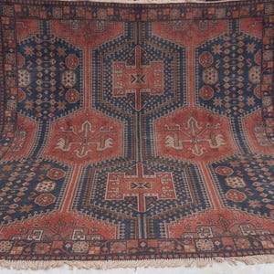 Pu&ograve; includere: Un grande tappeto rettangolare con un motivo geometrico dettagliato. Il tappeto presenta un disegno centrale nei toni del rosso e del marrone, incorniciato da un bordo blu scuro. Il motivo intricato include forme e simboli stilizzati. Il tappeto ha un bordo sfrangiato.