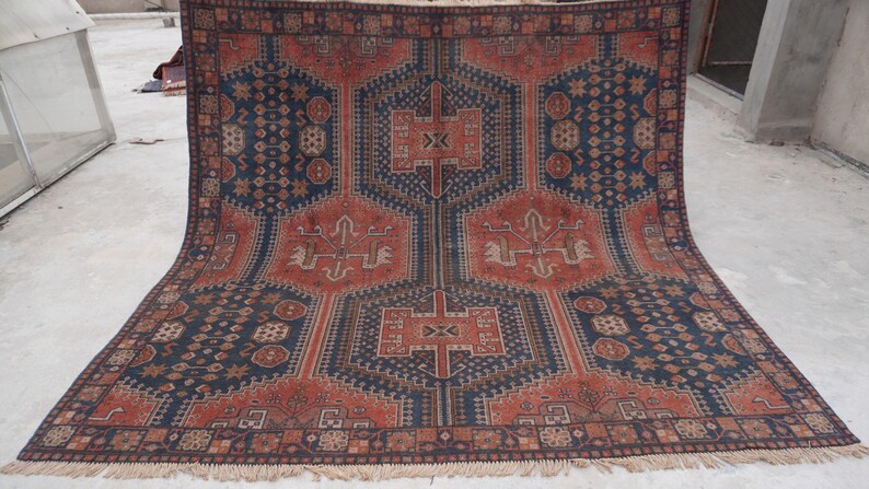 Pu&ograve; includere: Un grande tappeto rettangolare con un motivo geometrico dettagliato. Il tappeto presenta un disegno centrale nei toni del rosso e del marrone, incorniciato da un bordo blu scuro. Il motivo intricato include forme e simboli stilizzati. Il tappeto ha un bordo sfrangiato.