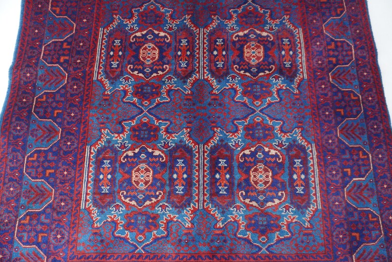 May include: A rectangular, patterned rug with a central design in shades of blue, red, and cream. The rug features an intricate, symmetrical pattern with floral and geometric elements. The border is a darker blue with repeating geometric shapes.