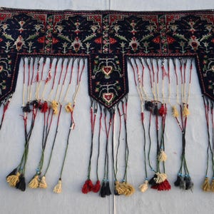 K&ouml;nnte beinhalten: Dunkelblaues Textil mit detailliertem Blumenmuster in Rot, Gr&uuml;n und Wei&szlig;. Das Textil hat einen Fransenboden mit Quasten in verschiedenen Farben. Das Design umfasst sich wiederholende Blumenmotive und dekorative Bord&uuml;ren.