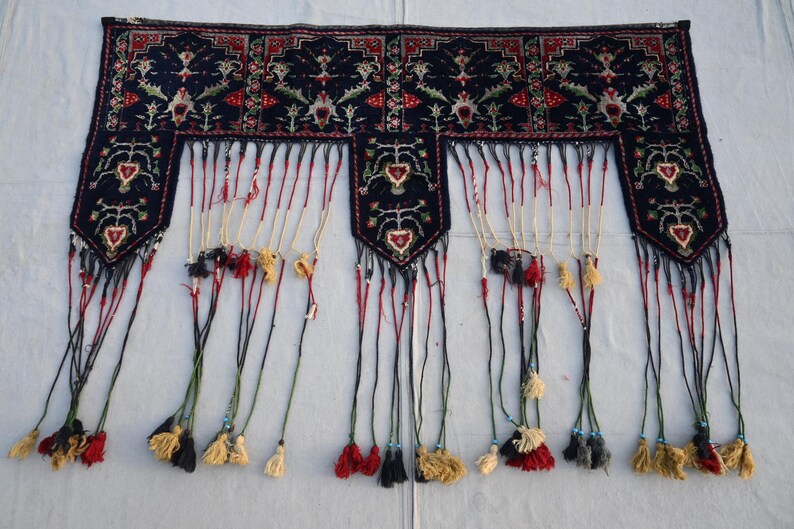 K&ouml;nnte beinhalten: Dunkelblaues Textil mit detailliertem Blumenmuster in Rot, Gr&uuml;n und Wei&szlig;. Das Textil hat einen Fransenboden mit Quasten in verschiedenen Farben. Das Design umfasst sich wiederholende Blumenmotive und dekorative Bord&uuml;ren.
