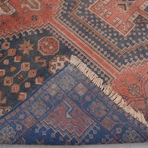 Pu&ograve; includere: Un tappeto antico con un motivo geometrico dettagliato nei toni blu navy, ruggine e beige. Il design del tappeto presenta forme ripetute e un bordo sfrangiato, mostrando un'estetica tradizionale.