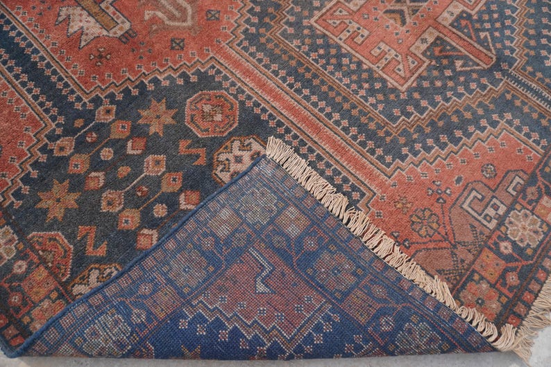 Pu&ograve; includere: Un tappeto antico con un motivo geometrico dettagliato nei toni blu navy, ruggine e beige. Il design del tappeto presenta forme ripetute e un bordo sfrangiato, mostrando un'estetica tradizionale.