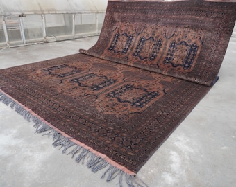 May include: A large, rectangular rug with an intricate, repeating pattern in shades of brown and navy blue. The rug has a rolled-up section, revealing the detailed design. The edges are finished with a fringe.