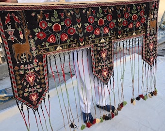 May include: A black textile with a deer and floral design, featuring red, green, and brown accents. The textile has a fringed bottom with tassels in various colors. The design is symmetrical and detailed.