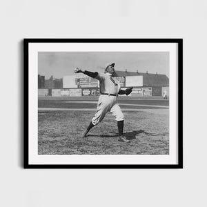 May include: Black and white framed print of a baseball player in action, captured mid-pitch. The player is wearing a baseball uniform, cap, and glove. The background shows a baseball field and buildings.