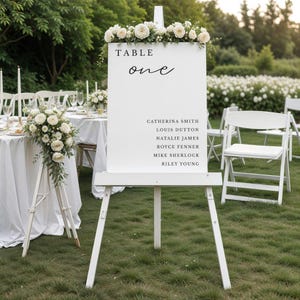 Op de afbeelding: Een witte ezel met een bord met de tekst "TABLE one" in zwart schrift. Het bord bevat namen en is versierd met witte bloemen. De scène speelt zich buiten af met witte stoelen en tafels.