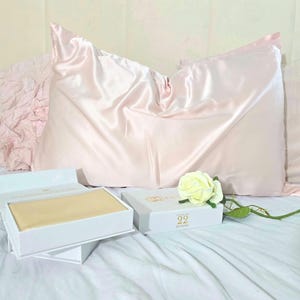 Op de afbeelding: Een lichtroze satijnen kussensloop op een bed met een wit hoofdbord. Een open witte doos met een goudkleurig item staat naast een gesloten doos met het nummer 22. Een witte roos ligt op het bed.