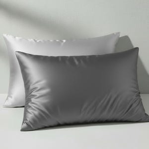 May include: Two satin pillowcases, one white and one gray, are displayed. The pillowcases have a smooth, shiny texture and a rectangular shape. The gray pillowcase is in the foreground, with the white pillowcase slightly behind it.