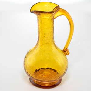 Puede incluir: Jarra de vidrio color ámbar con efecto craquelado. La jarra tiene un cuerpo redondeado, un pico y un asa curva. La base es de un color ámbar más oscuro. La jarra es probablemente vintage y adecuada para servir bebidas.
