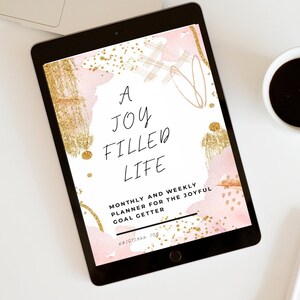 May include: A digital planner cover with a pink and gold watercolor background. The text reads "A Joy Filled Life Monthly and Weekly Planner for the Joyful Goal Getter" with the name "Kristiana Joy" at the bottom.