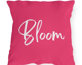 Pode incluir: Uma almofada quadrada fúcsia com a palavra "Bloom" escrita em branco, em estilo manuscrito. A almofada tem um fundo fúcsia liso, criando um design simples e elegante, adequado para a decoração da casa.
