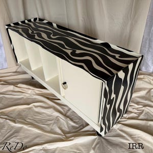 May include: A long, rectangular storage cabinet featuring a black and cream wavy, marbled pattern on the top and side panels. The front includes open shelving sections and a cabinet door with a circular silver handle, supported by small black legs.