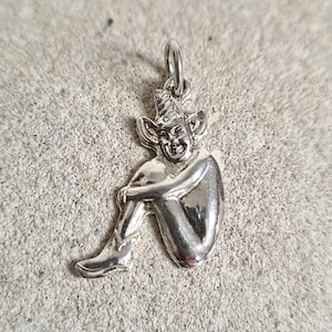 Puede incluir: Encanto de plata de una hada sentada con orejas puntiagudas y un sombrero. El hada se está sujetando las piernas y tiene una cara detallada.