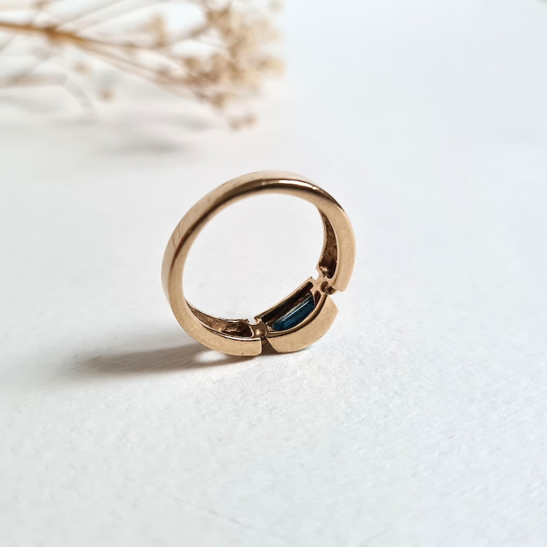 May include: A gold-colored ring with a rectangular, dark blue gemstone set in the center. The ring has a unique design with a split band. The ring is shown on a white surface.