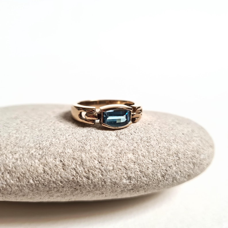 May include: A gold-toned ring with a rectangular, blue gemstone. The ring is set on a gray stone against a white background. The ring's design features a rectangular blue stone in the center, with decorative elements on either side.