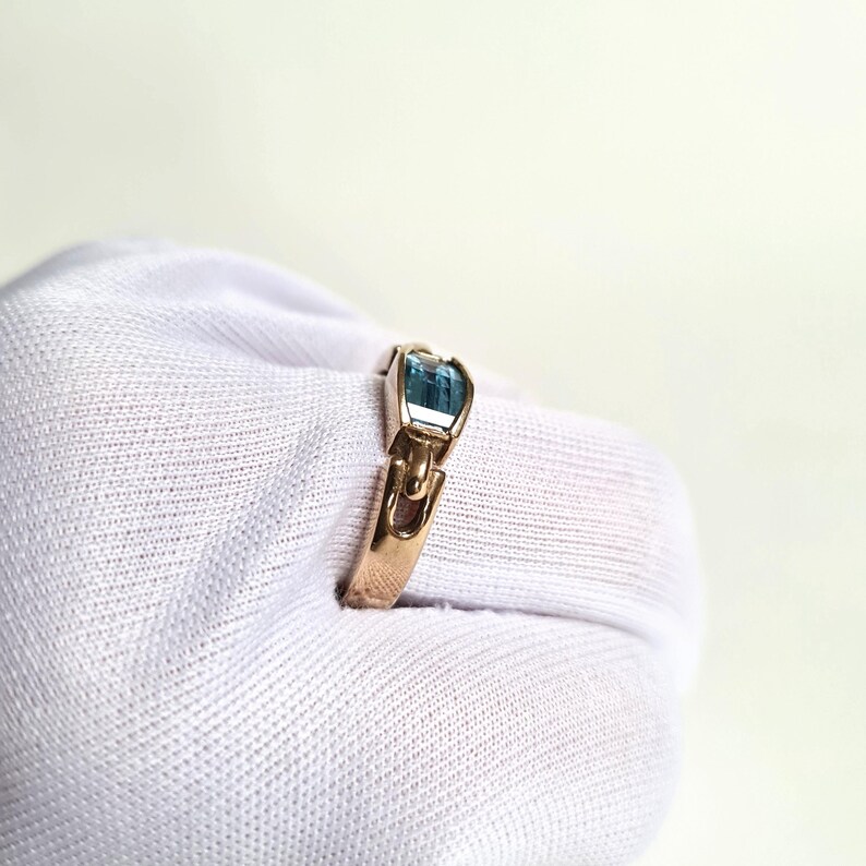 May include: A gold ring featuring a rectangular, light blue gemstone. The ring's band has a unique design with a decorative element on each side of the gemstone. The ring is displayed on a white surface.