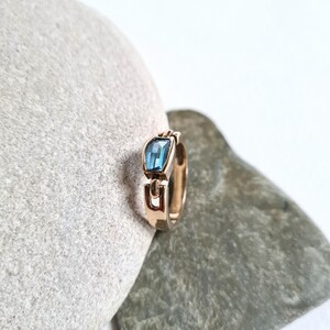 May include: A gold-toned ring with a rectangular, light blue gemstone. The ring's band features a unique, open design with a geometric shape. The gemstone is set in a bezel setting, and the ring is displayed on a gray stone surface.
