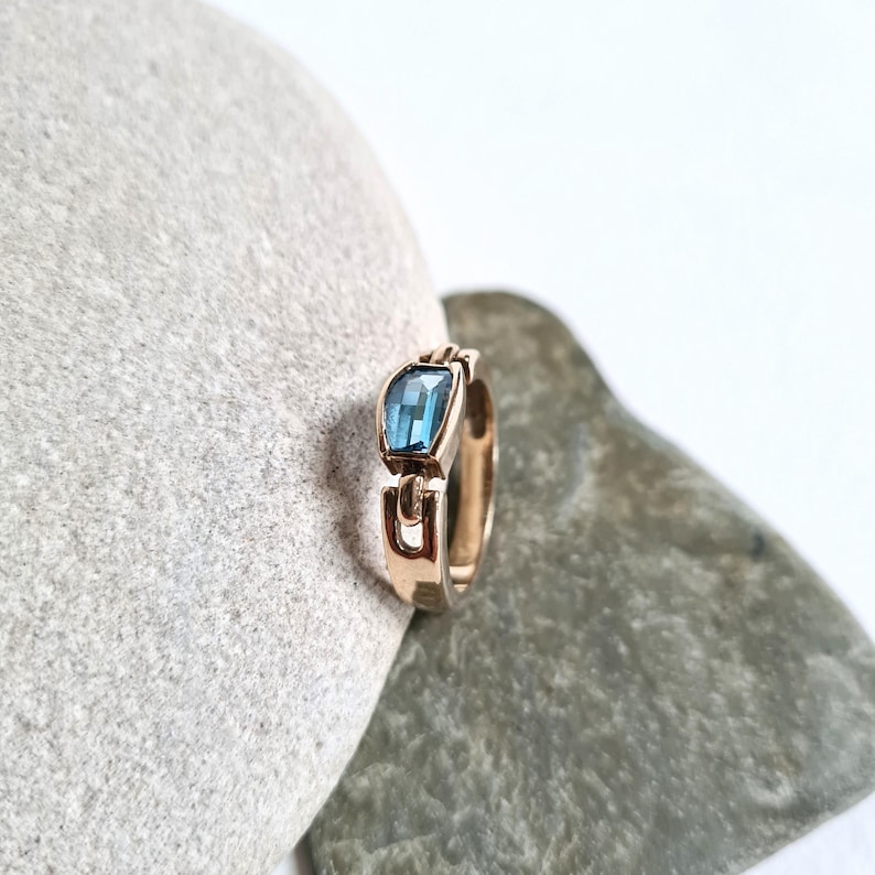 May include: A gold-toned ring with a rectangular, light blue gemstone. The ring's band features a unique, open design with a geometric shape. The gemstone is set in a bezel setting, and the ring is displayed on a gray stone surface.