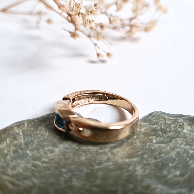 May include: A gold-toned ring with a rectangular blue gemstone. The ring is set on a gray stone, with dried flowers in the background. The ring's band has a smooth, polished finish.