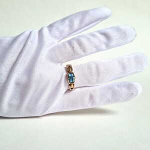 May include: A gold-colored ring with a rectangular, light blue gemstone. The ring is set on a white glove, showcasing the jewelry's design and color contrast. The gemstone is approximately 0.3 inches in length.
