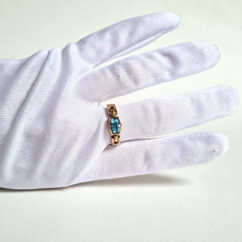May include: A gold-colored ring with a rectangular, light blue gemstone. The ring is set on a white glove, showcasing the jewelry's design and color contrast. The gemstone is approximately 0.3 inches in length.