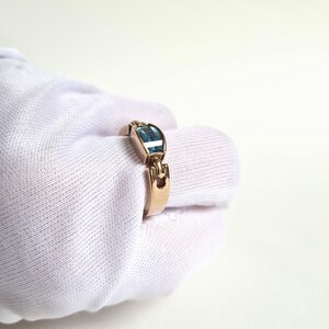 May include: A gold-toned ring featuring a rectangular, light blue gemstone. The ring has a unique design with a decorative element on the band. The ring is shown on a white background.