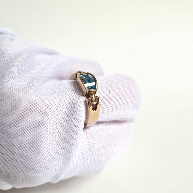 May include: A gold-toned ring featuring a rectangular, light blue gemstone. The ring has a unique design with a decorative element on the band. The ring is shown on a white background.