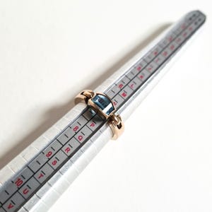 May include: A gold-colored ring with a rectangular, light blue gemstone. The ring is on a silver ring sizer. The ring is a size 8.