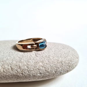 May include: A gold-colored ring with a rectangular, light blue gemstone. The ring is set on a gray stone against a white background. The ring's design features a unique clasp-like detail on the side.