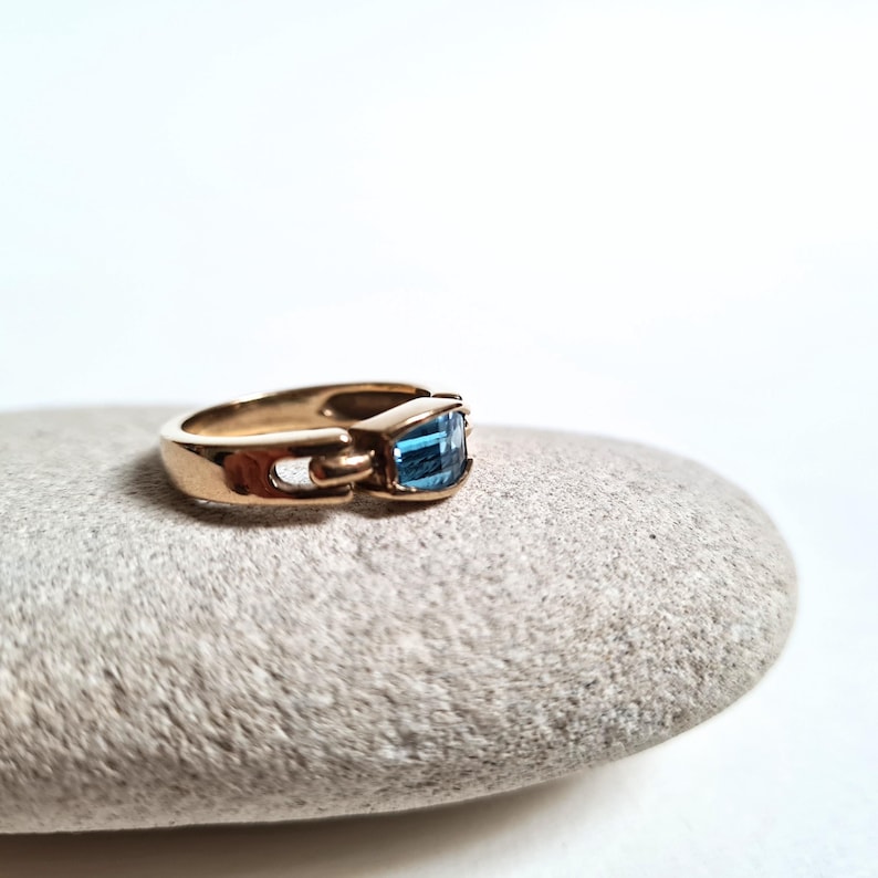 May include: A gold-colored ring with a rectangular, light blue gemstone. The ring is set on a gray stone against a white background. The ring's design features a unique clasp-like detail on the side.
