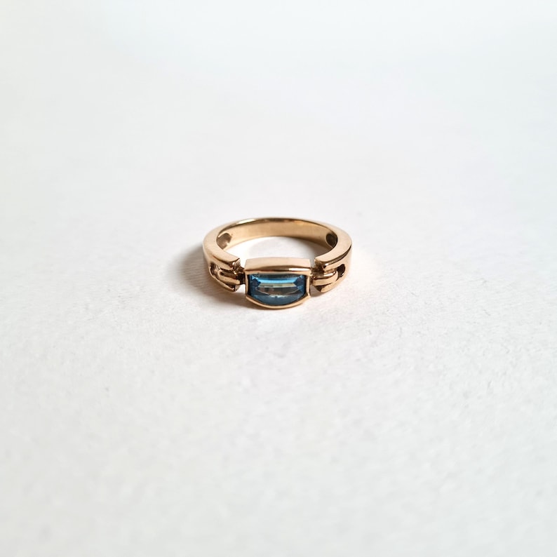 May include: A gold ring with a rectangular, light blue gemstone set in the center. The ring has a simple, elegant design with a polished finish. The band is a warm gold color, and the gemstone is a cool, contrasting blue.