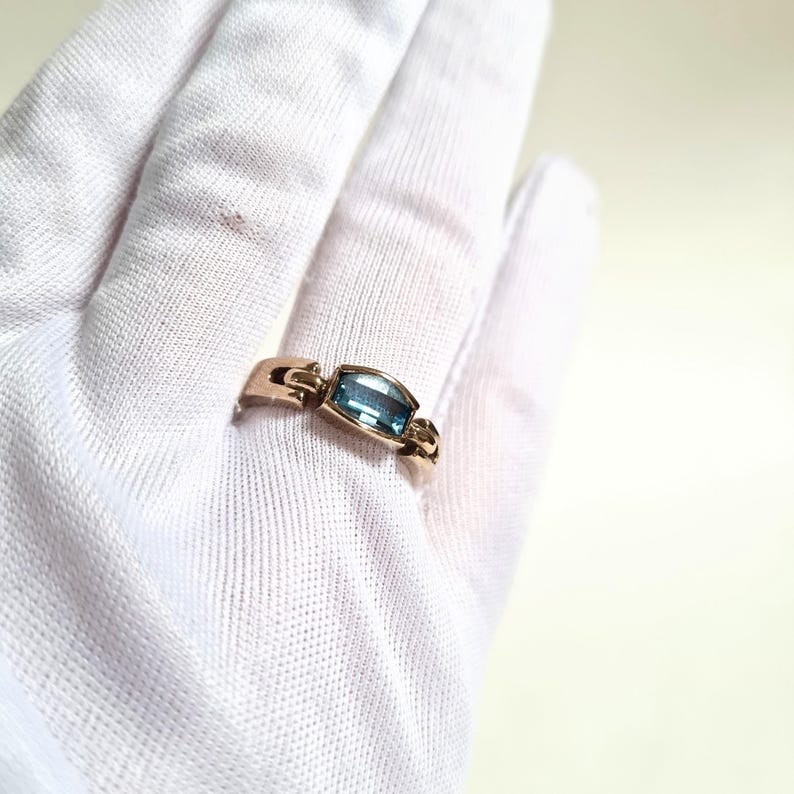 May include: A gold-toned ring featuring a rectangular, light blue gemstone. The ring's band has a unique design with a central setting for the gemstone. The ring is displayed on a white surface.