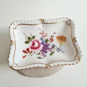 May include: A rectangular, white porcelain dish with a scalloped edge and gold trim. The dish is decorated with a floral pattern featuring pink roses, purple and blue flowers, and an orange daisy. The dish is resting on a gray, textured surface.